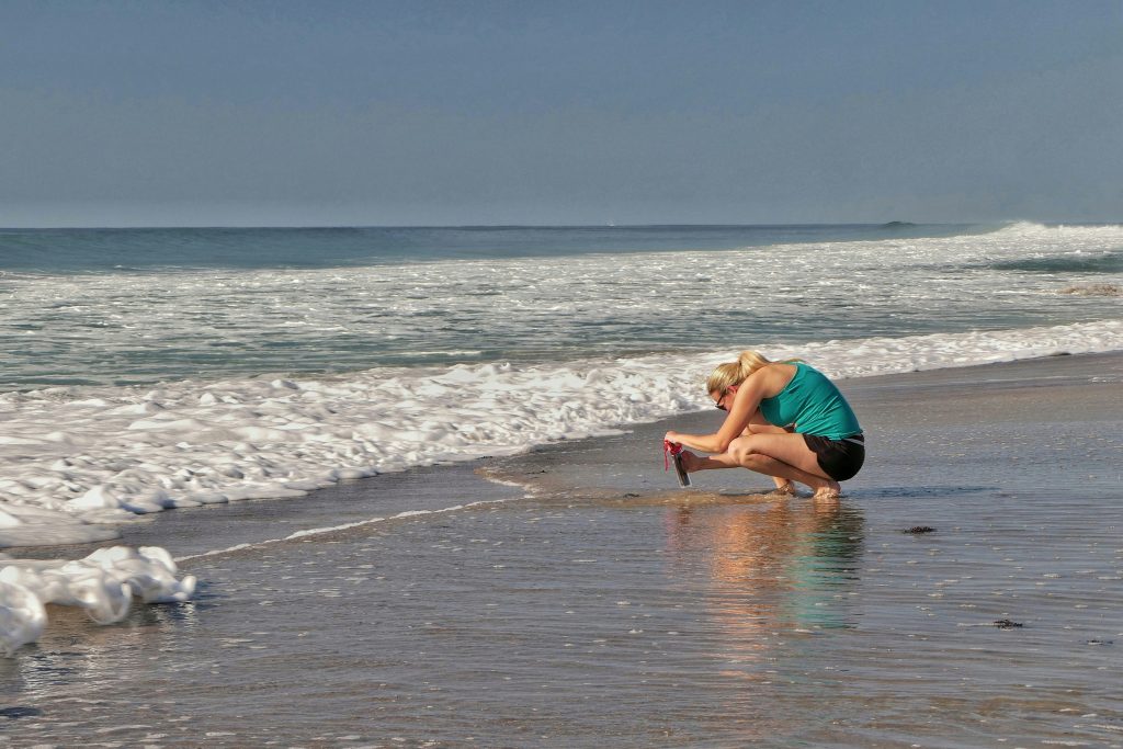 Blonde Frau, die am Strand hockt mit dem Handy die Wellen filmt.